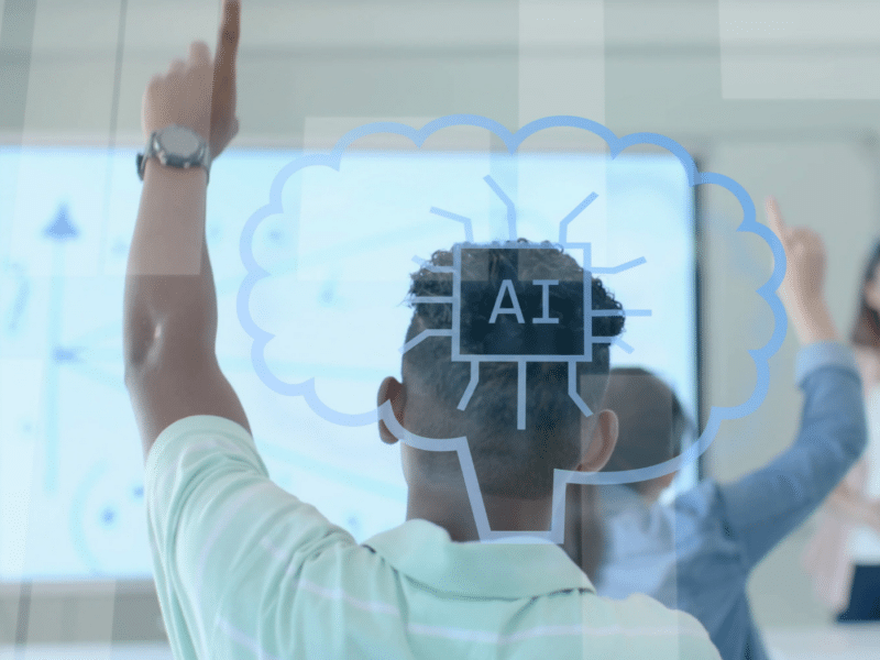 A mixed raced teen boy has back turned to the camera, with his left hand raised in the area. He is wearing a light mint green polo. He appears to be in a learning environment, like a school. In between the camera and the room is a glass pane that has a speech mark icon in a light gray colour, with "AI" in the middle.