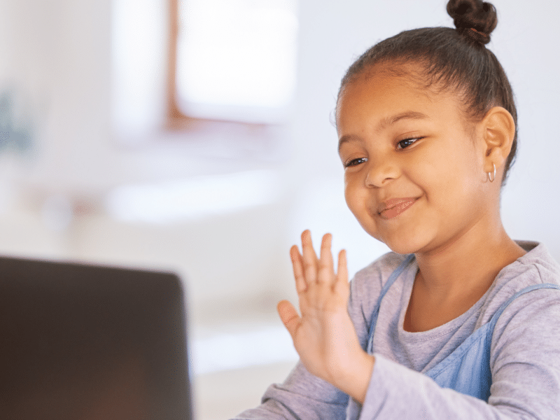 A young black girl on a laptop. She is smiling and waves at the laptop in front of her, which suggests she is on a video call.