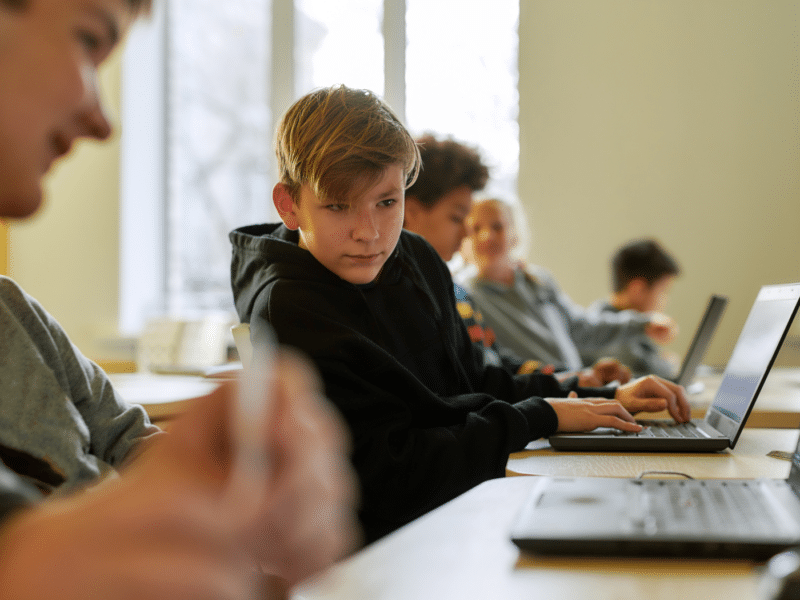 Schoolboy looking at the laptop of his classmate while sitting in a classroom with other pupils during a lesson. His classmate's face is in the top left corner in the foreground of the image but blurred, as the camera is focused on the schoolboy looking at his laptop.