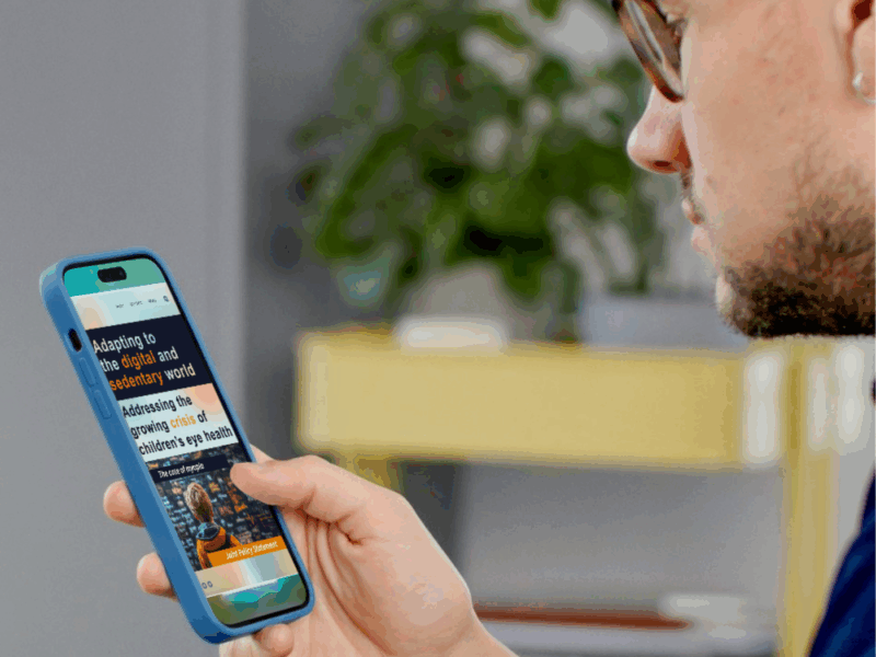 A man wearing glasses and a navy shirt is holding a smartphone in his left hand, closely reading an article. The phone has a blue case, and the screen displays a webpage titled 'Adapting to the digital and sedentary world' with a subheading about the crisis in children's eye health.