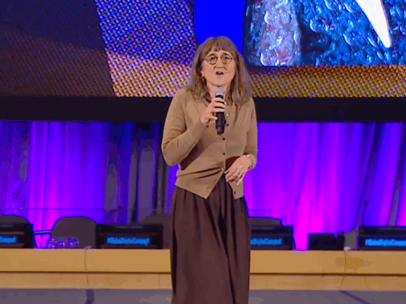 Baroness Beeban Kidron stands on a stage at the UN STI Forum, holding a microphone while addressing the audience. She wears a brown cardigan and long skirt. This is a wide shot of the picture shown above.