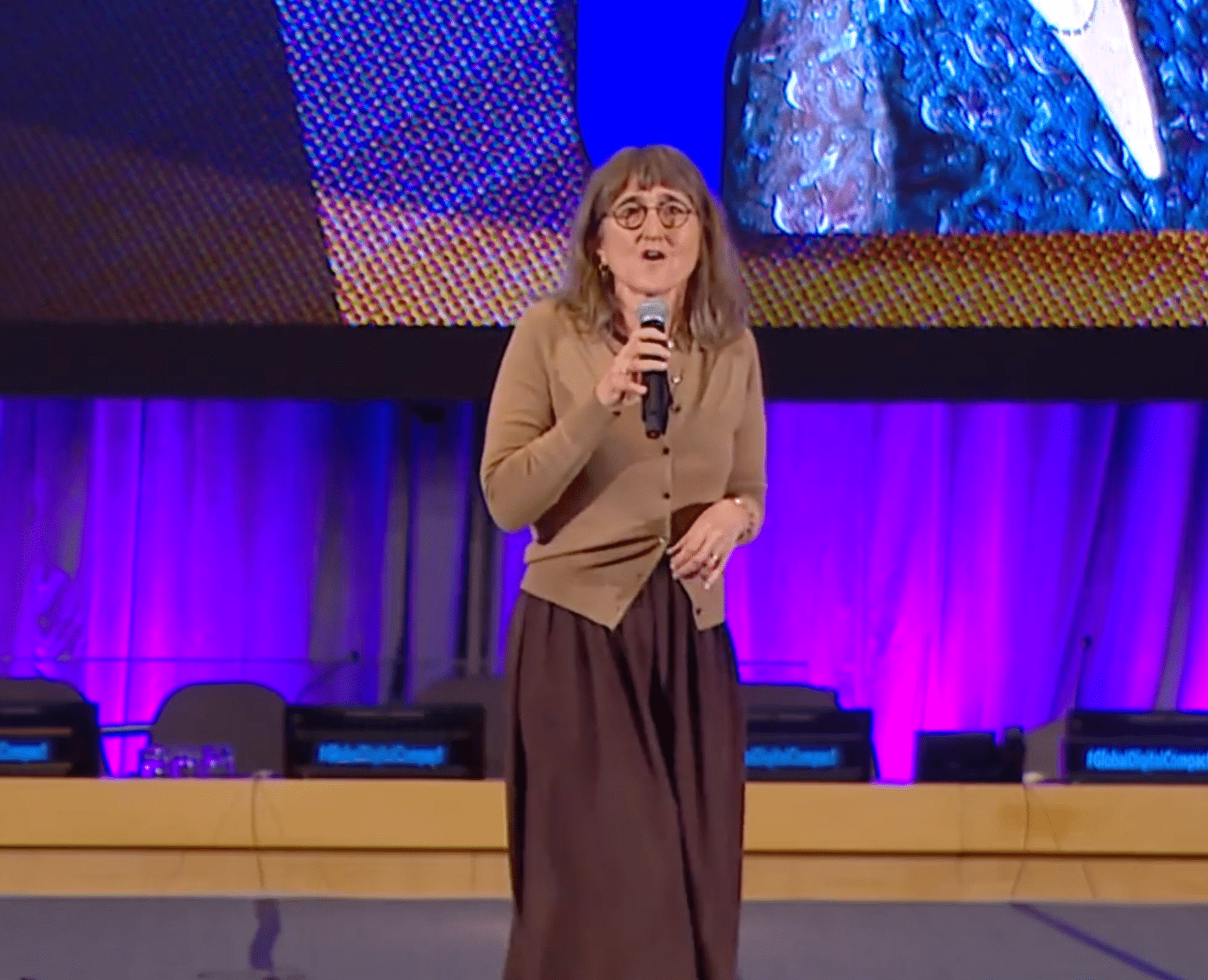 Baroness Beeban Kidron stands on a stage at the UN STI Forum, holding a microphone while addressing the audience. She wears a brown cardigan and long skirt. This is a wide shot of the picture shown above.
