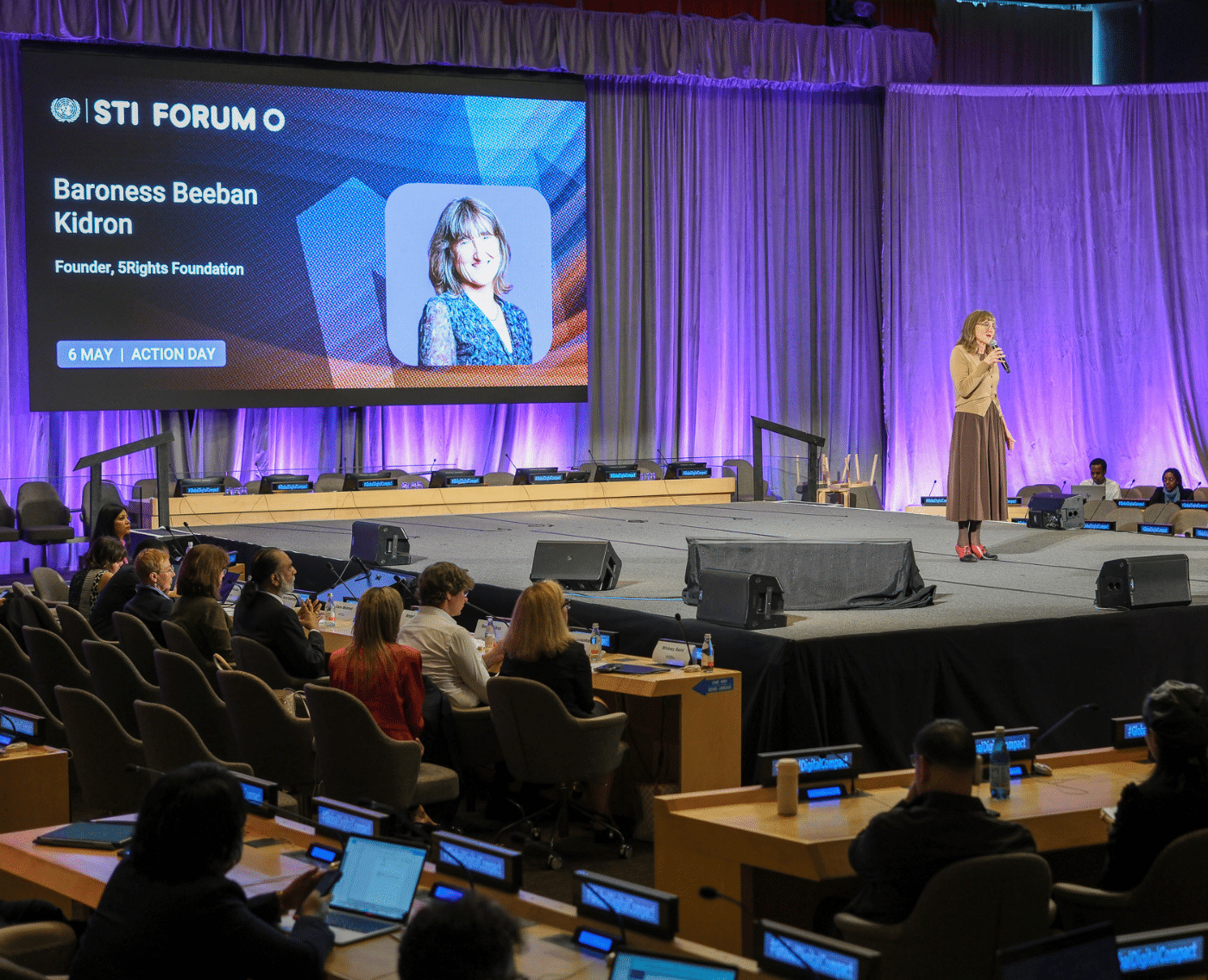 Baroness Beeban Kidron stands on a stage at the UN STI Forum, holding a microphone while addressing the audience. She wears a brown cardigan and long skirt. This is a wide shot of the picture shown above.