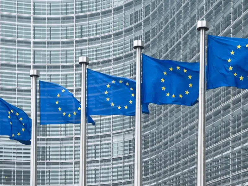 A wider view showing a row of European Union flags in front of a modern, glass-fronted office building with a curved facade. The flags are aligned in front of the European Commission headquarters in Brussels.