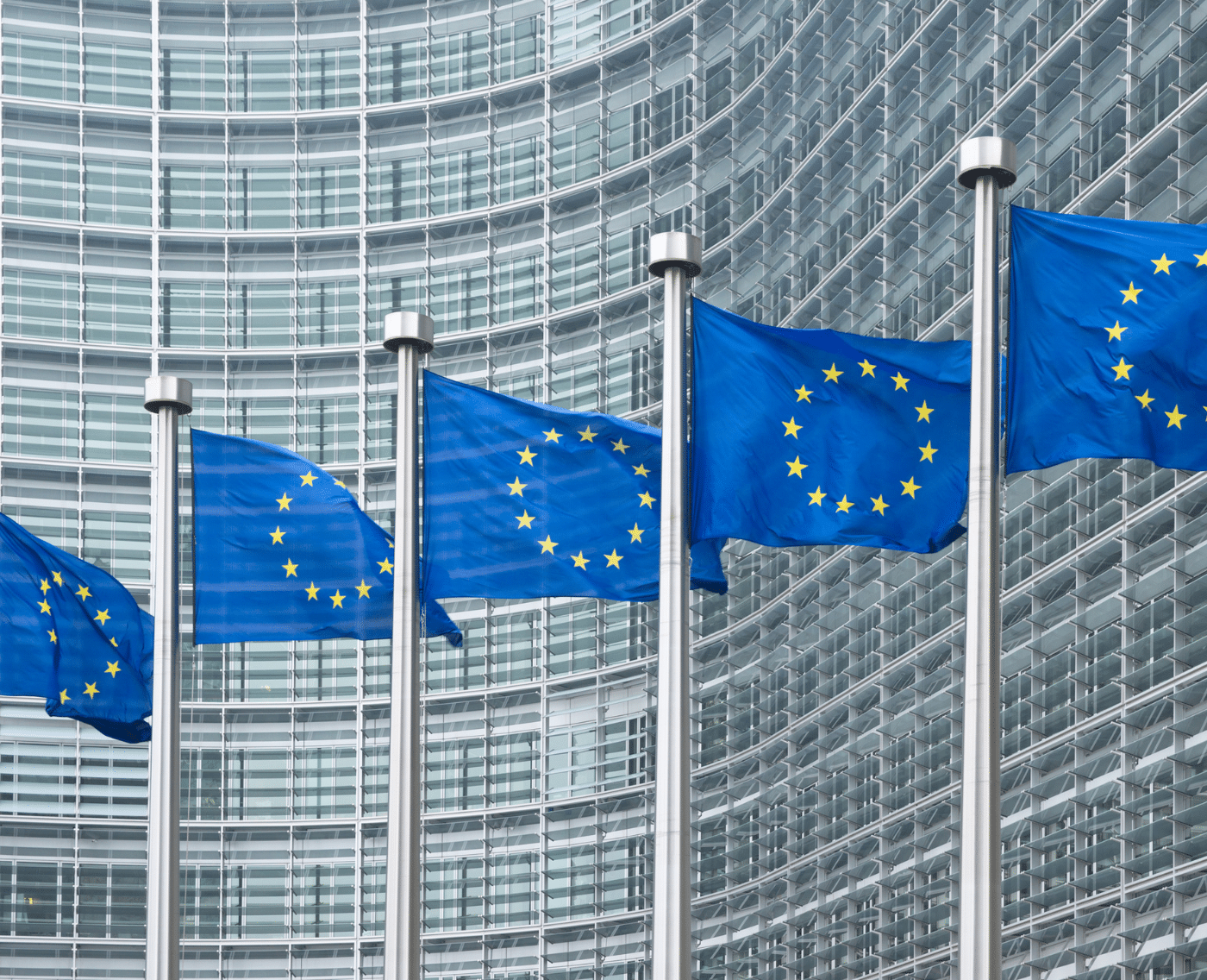 A wider view showing a row of European Union flags in front of a modern, glass-fronted office building with a curved facade. The flags are aligned in front of the European Commission headquarters in Brussels.