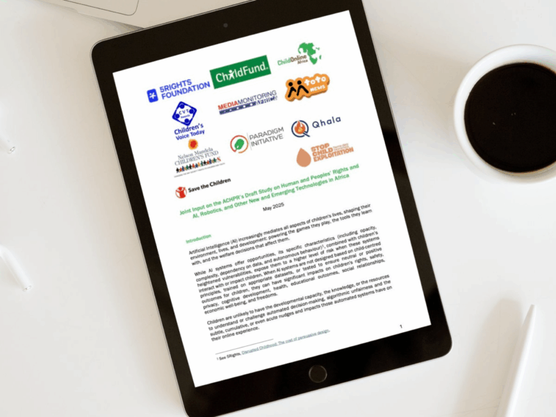 A tablet on a white desk displays a document titled "Joint Input on the ACHPR’s Draft Study on Human and Peoples’ Rights and AI, Robotics, and Other New and Emerging Technologies in Africa”. The document is dated May 2025 and includes logos of several child rights organisations, such as 5Rights Foundation, ChildFund, Save the Children, Paradigm Initiative, and others. A cup of black coffee and a pair of glasses are also visible on the desk beside the tablet.