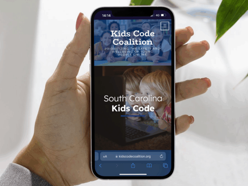 A woman holding a smartphone with an open palm. Her smartphone is on the South Carolina Kids Code website.
