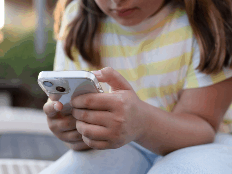 A wider shot showing a child in a yellow-and-white striped shirt and light blue jeans sitting outdoors, intently looking at and using a smartphone with both hands.