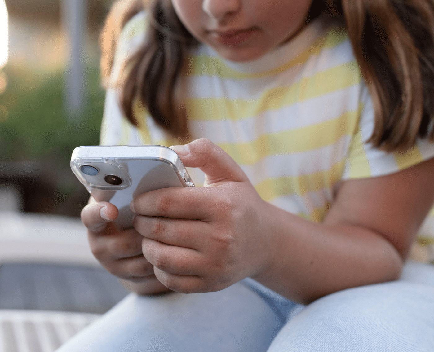 A wider shot showing a child in a yellow-and-white striped shirt and light blue jeans sitting outdoors, intently looking at and using a smartphone with both hands.