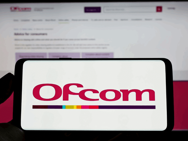 A siloheutte of a hand holding a smartphone horizontally in the foreground. It displays red text, 'Ofcom'. In the background, an out-of-focus monitor displays the Ofcom webiste.