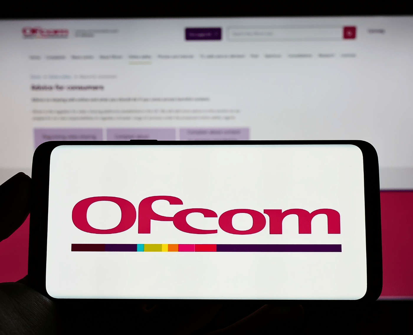 A siloheutte of a hand holding a smartphone horizontally in the foreground. It displays red text, 'Ofcom'. In the background, an out-of-focus monitor displays the Ofcom webiste.
