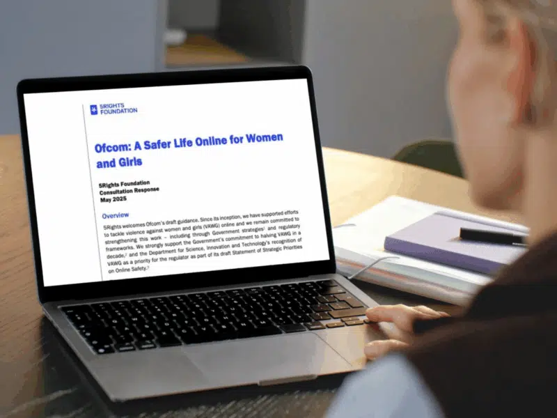 A person sits at a desk, viewing a laptop screen that displays a document titled "Ofcom: A Safer Life Online for Women and Girls," produced by the 5Rights Foundation. The document is a consultation response dated May 2025. The person’s face is partially visible, with focus on the screen. A stack of notebooks and a pen lie next to the laptop on the desk