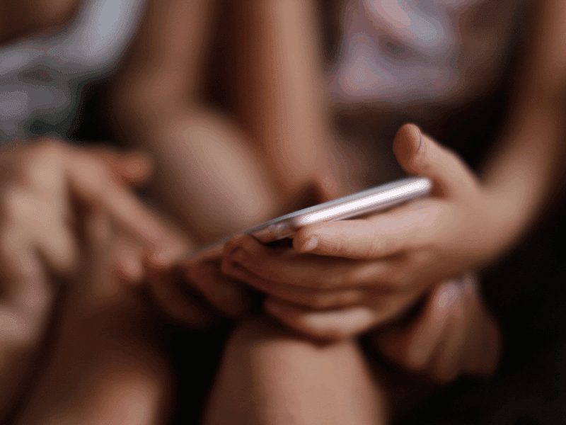 A child sits with a smartphone in their hands while another child points at the screen. The focus is on the interaction with the device.