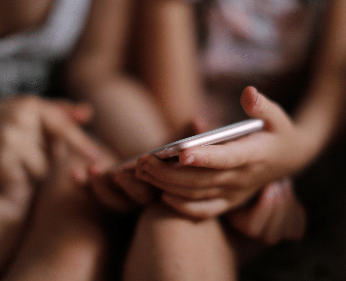 A child sits with a smartphone in their hands while another child points at the screen. The focus is on the interaction with the device. 