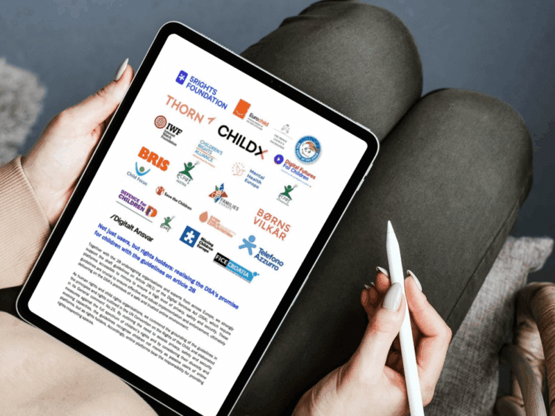 A person holding a tablet in their lap is reviewing a document displaying logos of various child rights and advocacy organisations, including 5Rights Foundation, Thorn and Save the Children, among others. The document is titled "Not just users, but rights holders: realising the DSA's promise for children with the guidelines on article 28," with a heading section featuring the logos of all the signatories of this joint statement.