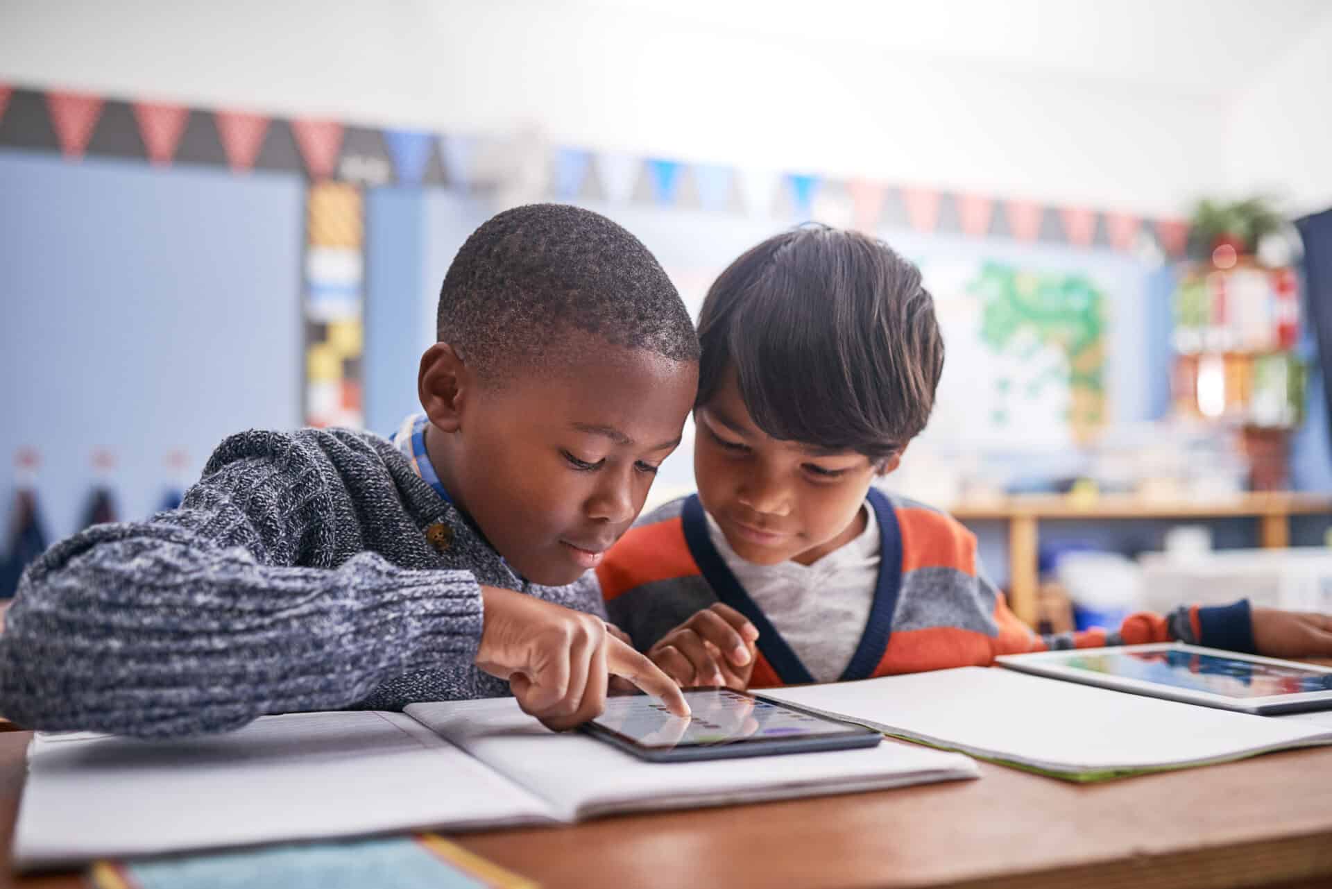Two elementary school students work together on a tablet in a colorful classroom. One child wears a dark gray sweater while the other wears an orange and gray color-block shirt. They're focused on the tablet screen at their desk, with books and papers nearby. In the background, decorations and educational displays create a bright, engaging learning environment.