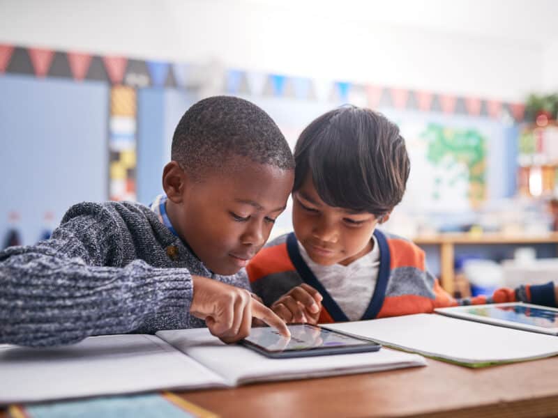 Two elementary school students work together on a tablet in a colorful classroom. One child wears a dark gray sweater while the other wears an orange and gray color-block shirt. They're focused on the tablet screen at their desk, with books and papers nearby. In the background, decorations and educational displays create a bright, engaging learning environment.