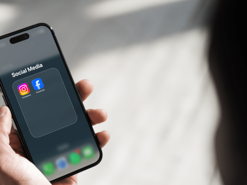 A person holds a smartphone displaying a "Social Media" folder containing the Instagram and Facebook apps, with the home screen dock visible below.