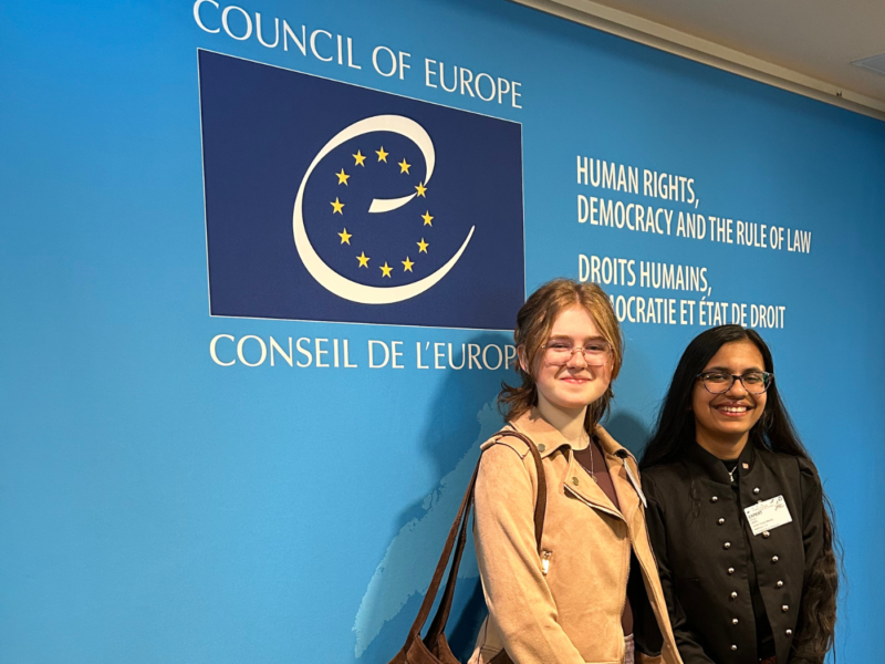 5Rights Youth Ambassador Rhythm, 17, from Malta (right), and 5Rights Youth Council member Julia, 16, from Ireland (left) are smiling in front of a blue wall with the logo of the Council of Europe.