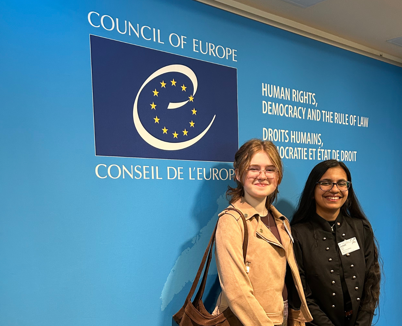5Rights Youth Ambassador Rhythm, 17, from Malta (right), and 5Rights Youth Council member Julia, 16, from Ireland (left) are smiling in front of a blue wall with the logo of the Council of Europe.