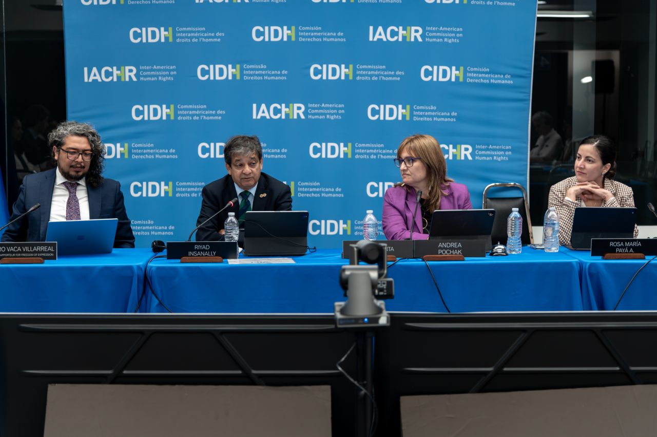 Four IACHR (CIDH) commissioners sit at a table during a hearing on accountability of digital platforms, in front of a blue backdrop with repeated CIDH/IACHR logos. Name plates identify them as (from left) Vaca Villarreal, Riyad Insanally, Andrea Pochak, and Rosa María Payá A., each with a laptop and water bottle in front of them. A video camera is visible in the foreground, framing the panel.